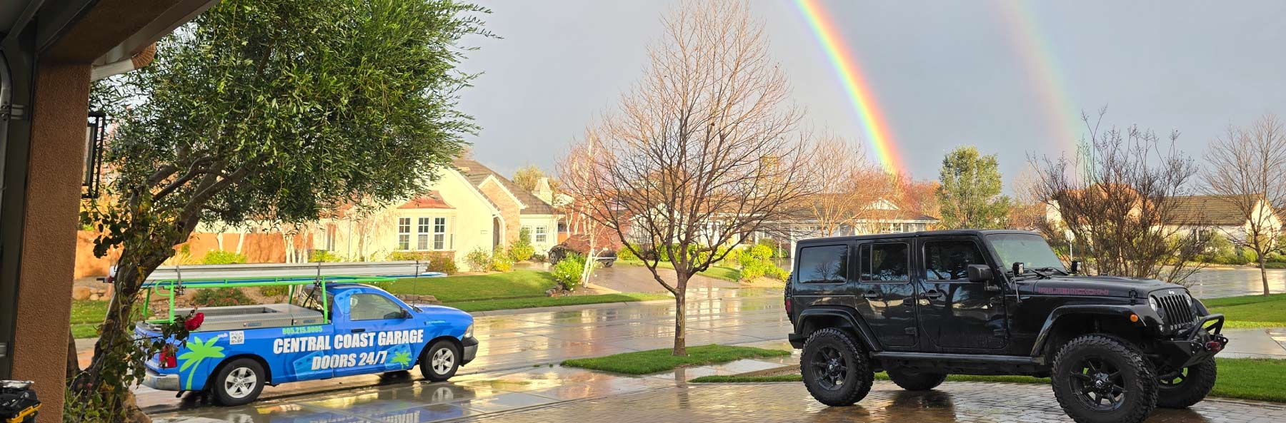 A blue Central Coast Garage Doors 24/7 service truck parked in a residential driveway in San Luis Obispo, CA, under a vibrant double rainbow. The branded mobile unit is equipped with professional ladders and equipment, ready to provide 24/7 emergency garage door repair to local homeowners