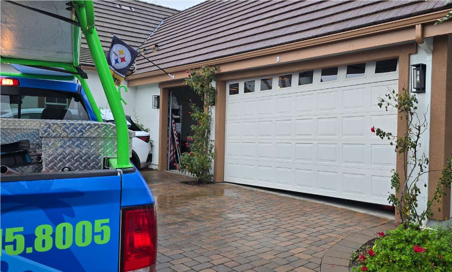 A professional technician from Central Coast Garage Doors 24/7 in San Luis Obispo, CA, expertly carrying two heavy-duty replacement springs during a residential repair job. A branded blue service truck with the company’s 24/7 contact information and license #1049595 is parked on the driveway against a scenic backdrop of the Central Coast hills
