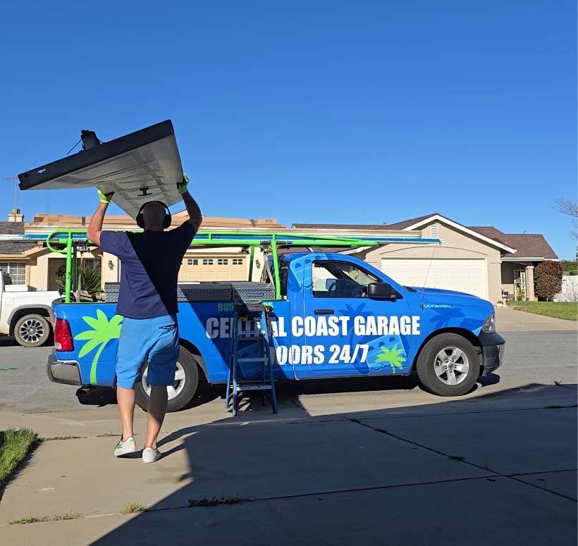 Central Coast Garage Doors 24/7 owner Cory Lewis carries a large garage door panel above his head toward a company truck, providing expert garage door repair in Atascadero for a local residence.