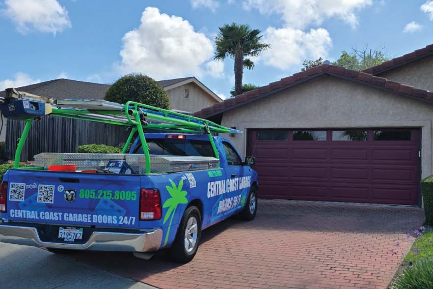 A blue Central Coast Garage Doors 24/7 service truck with green ladder racks parked on a brick driveway in front of a residential home with a closed purple-brown garage door.